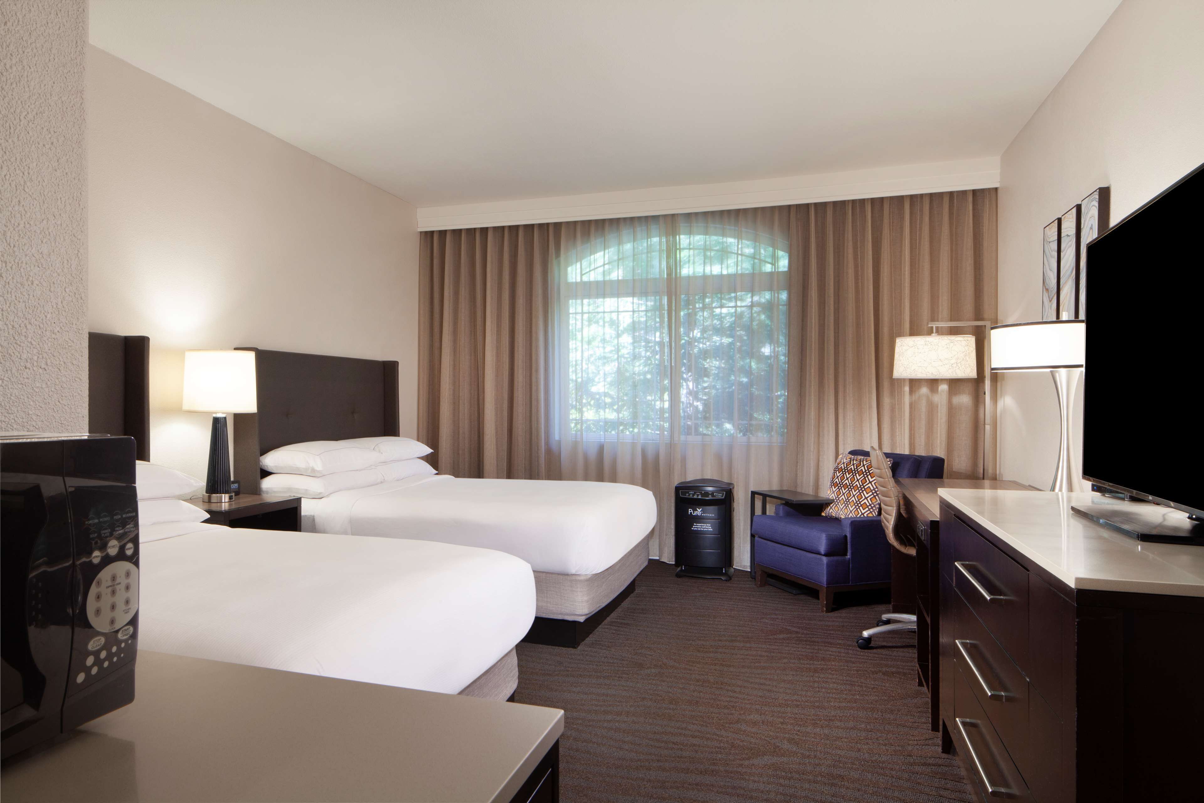 A hotel room with two beds, a desk, a TV, a lamp, and a window with curtains.