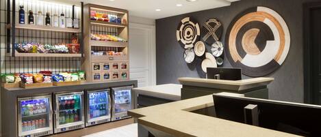 A modern reception area with a counter, refrigerated beverage dispensers, snack shelves, and decorative wall art.