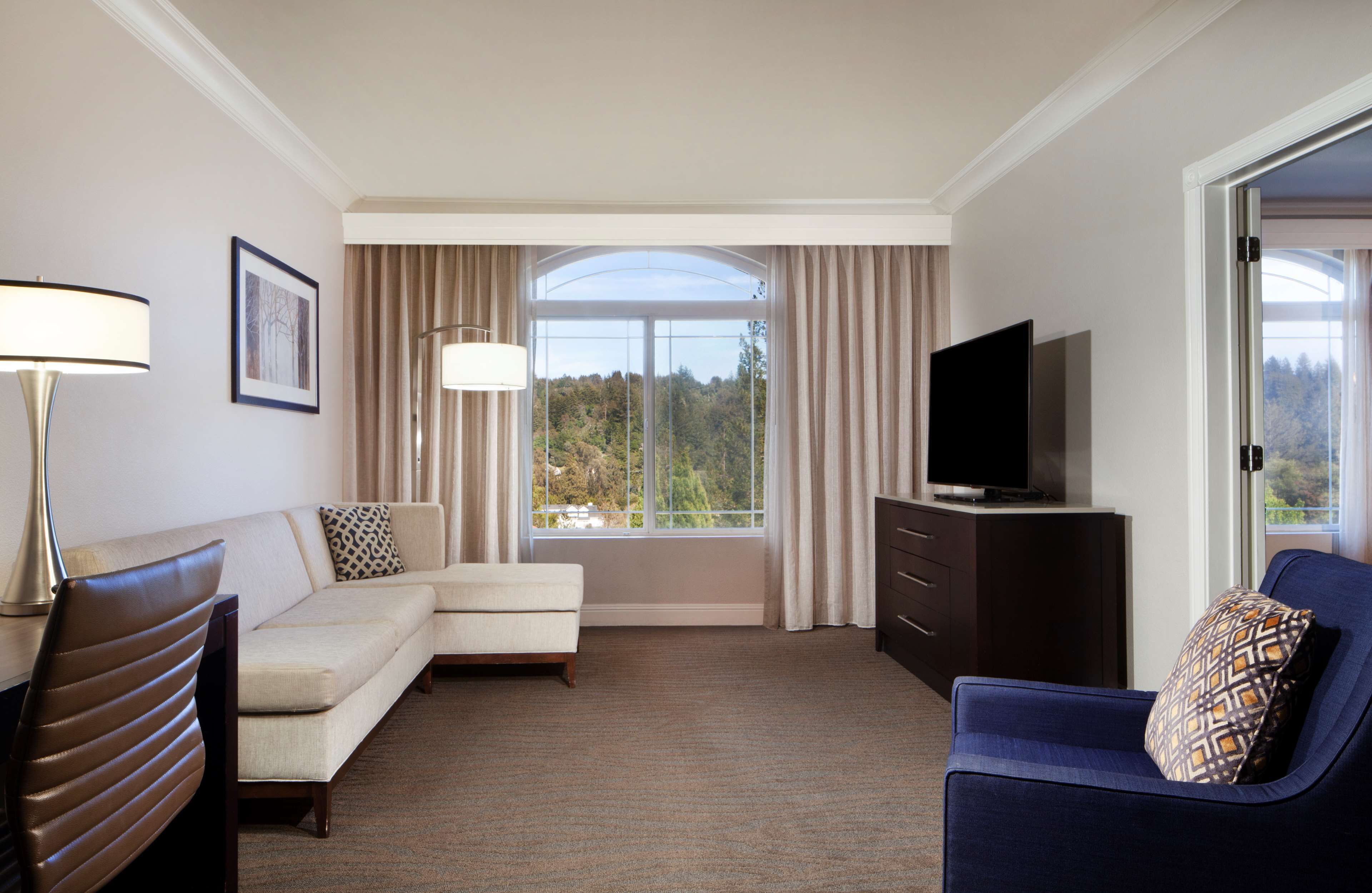 A hotel room with a sofa, a blue armchair, a TV on a dresser, and a large window with curtains.