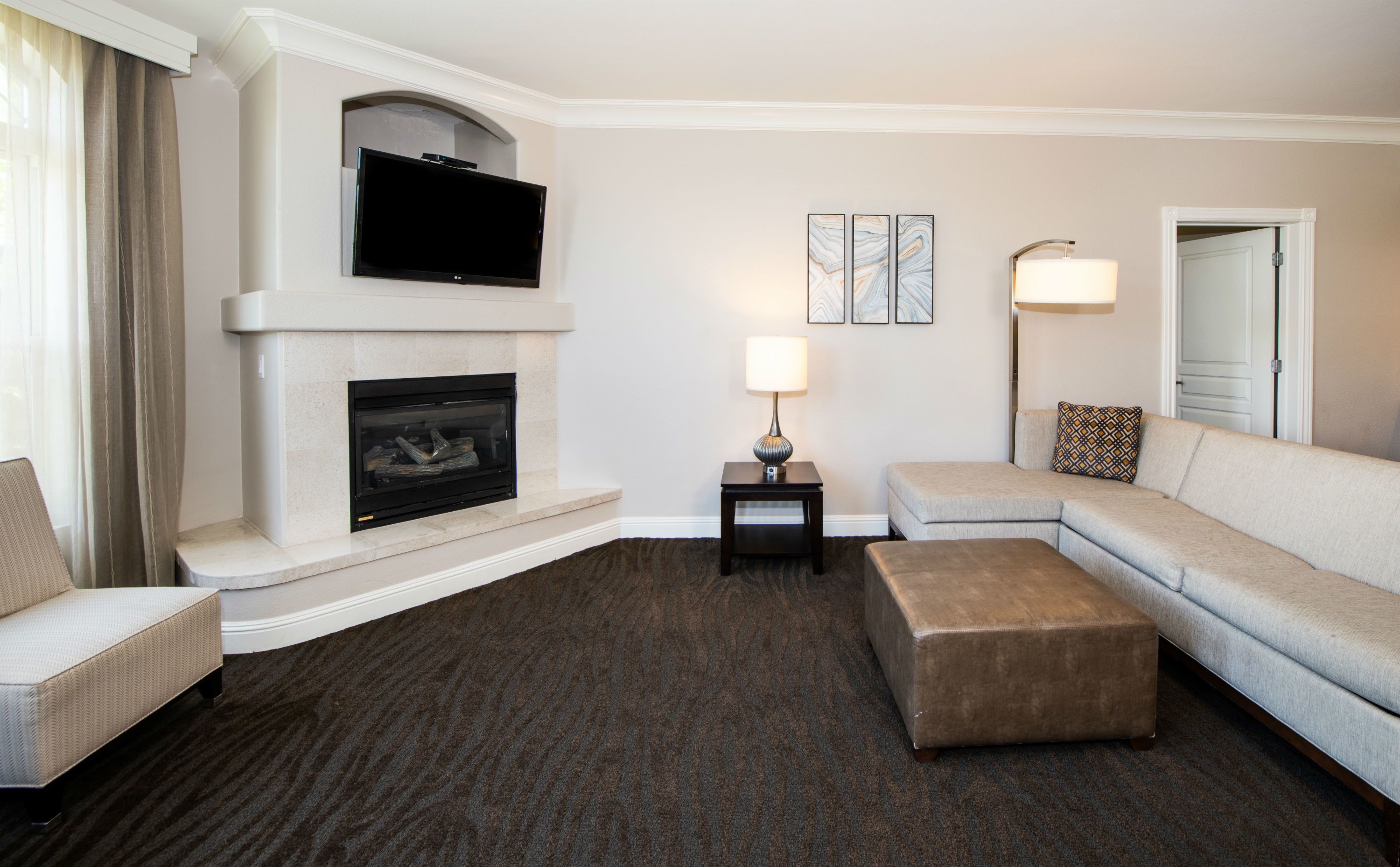 A modern living room with a fireplace, a flat-screen TV mounted above it, a sofa, an ottoman, a side table with a lamp, and a door leading to another room.