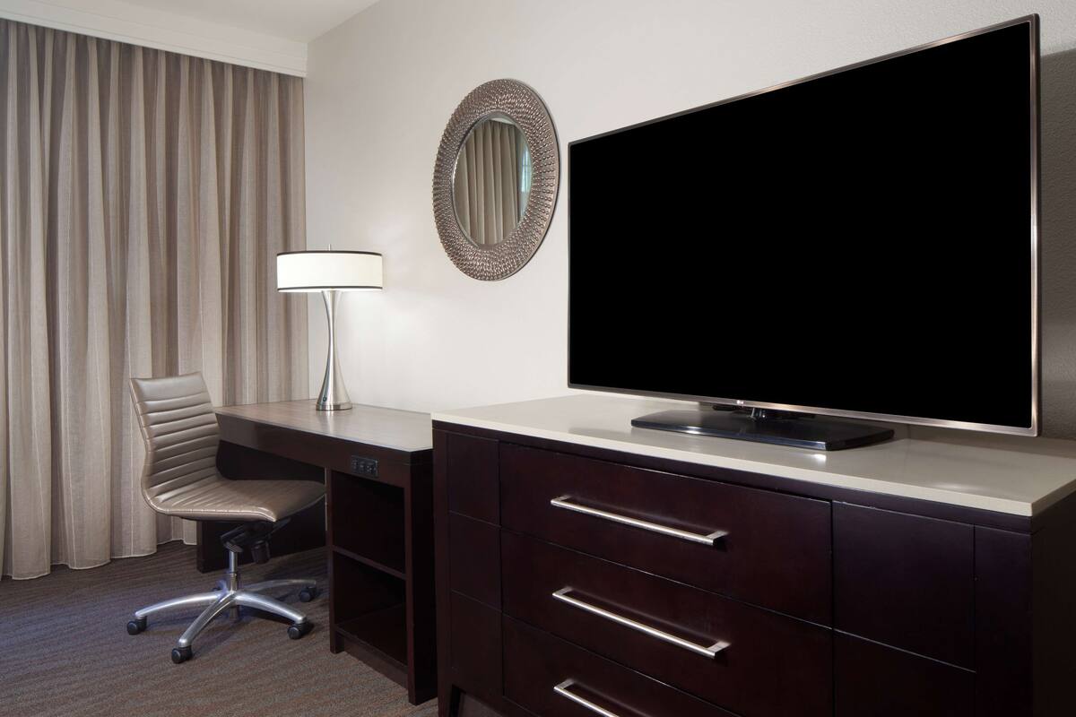Ein Hotelzimmer mit einem großen Flachbildfernseher, einem Schreibtisch mit Stuhl, einer Lampe und einer dunkelbraunen Kommode mit Schubladen.