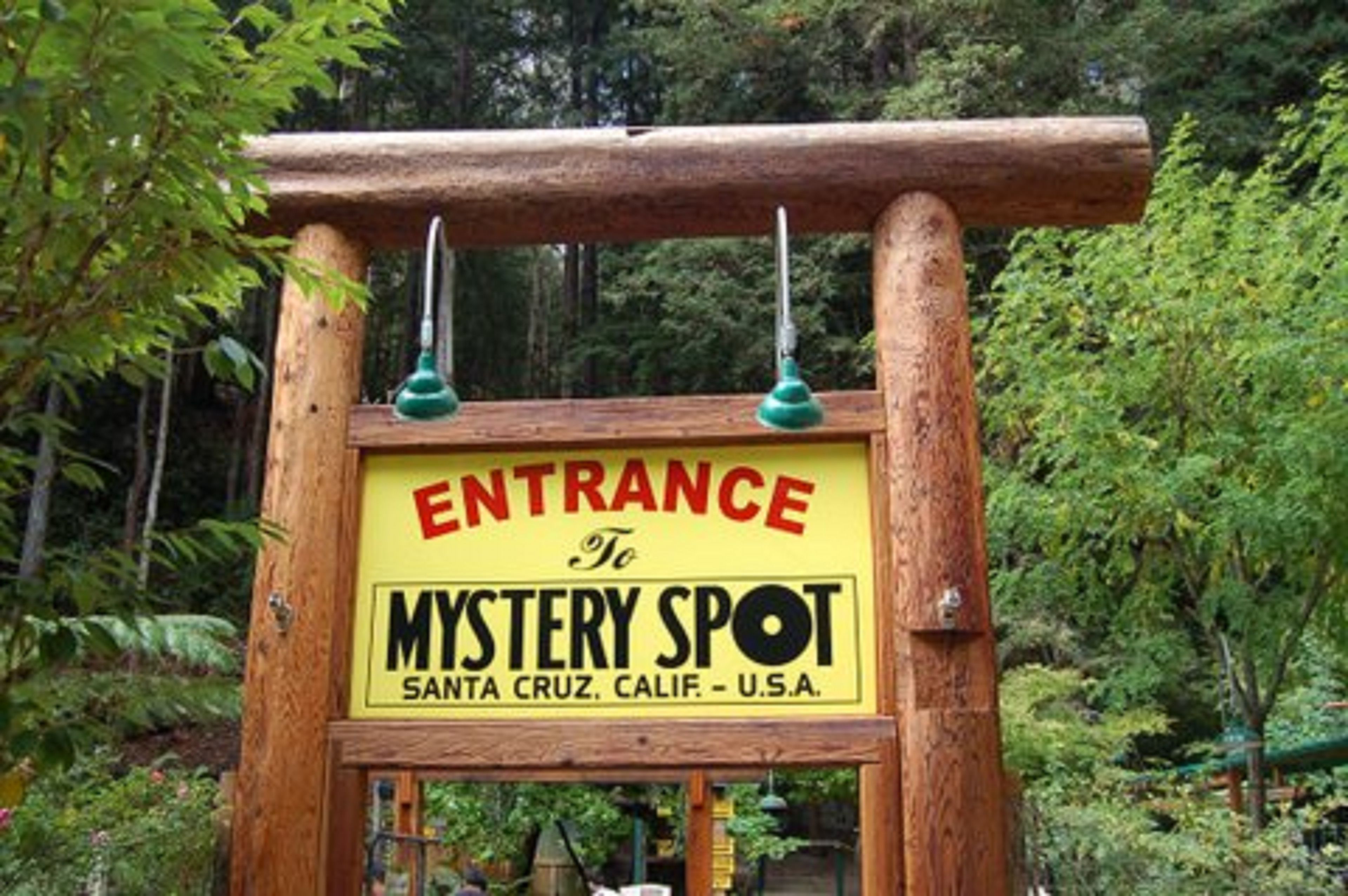 A wooden sign with a yellow background and black lettering that reads "ENTRANCE To MYSTERY SPOT Santa Cruz, Calif. - U.S.A."