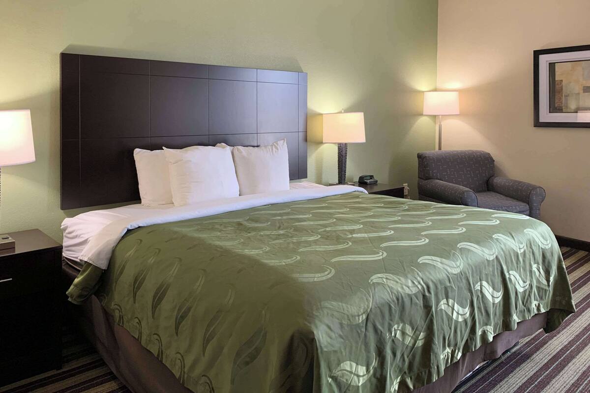 A hotel room with a large bed, a dark headboard, two bedside lamps, a chair, and a framed picture on the wall.