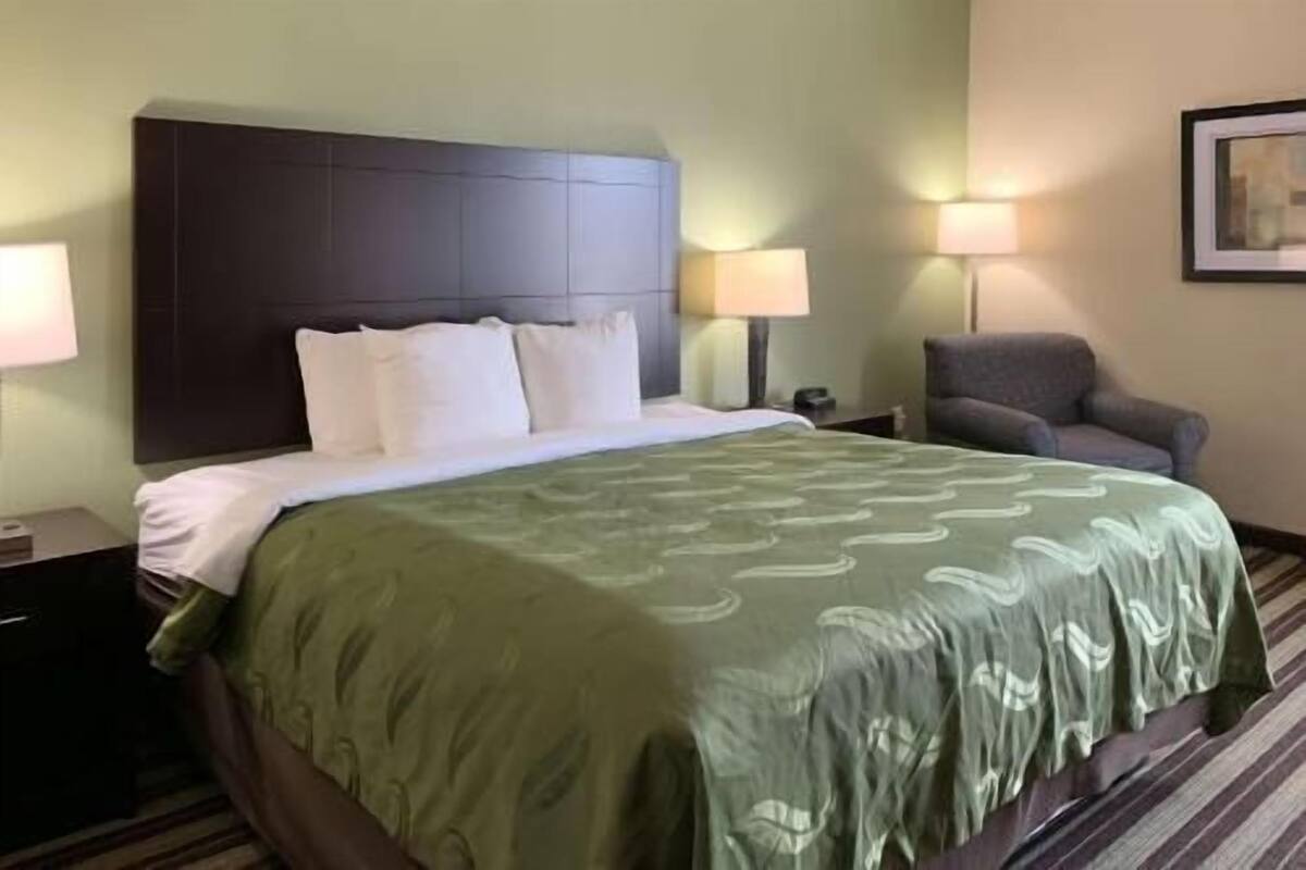 A hotel room with a large bed, a headboard, two bedside lamps, a chair, and a framed picture on the wall.