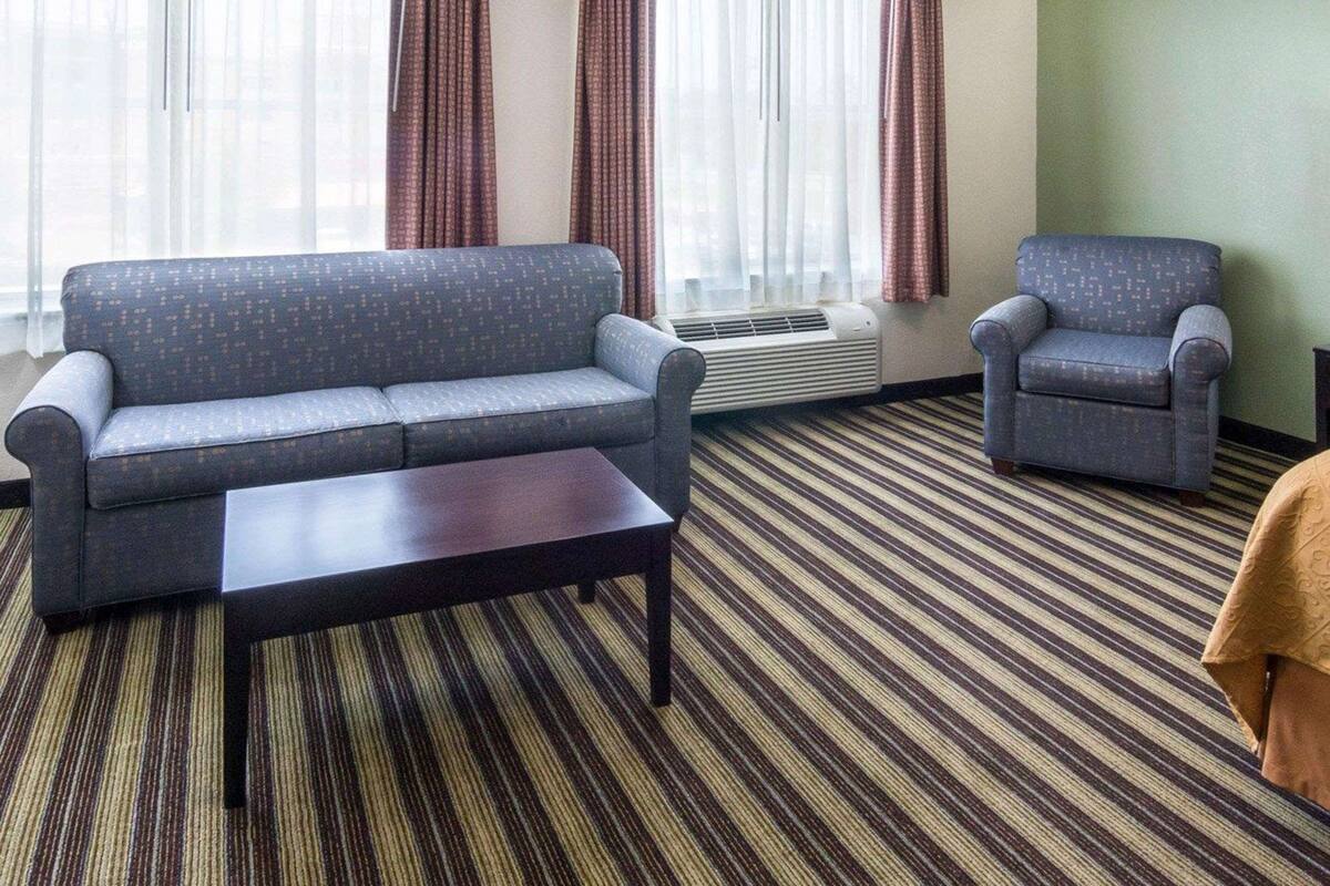 A hotel room with a sofa, armchair, and a small table. There is a window with curtains and an air conditioning unit on the wall.