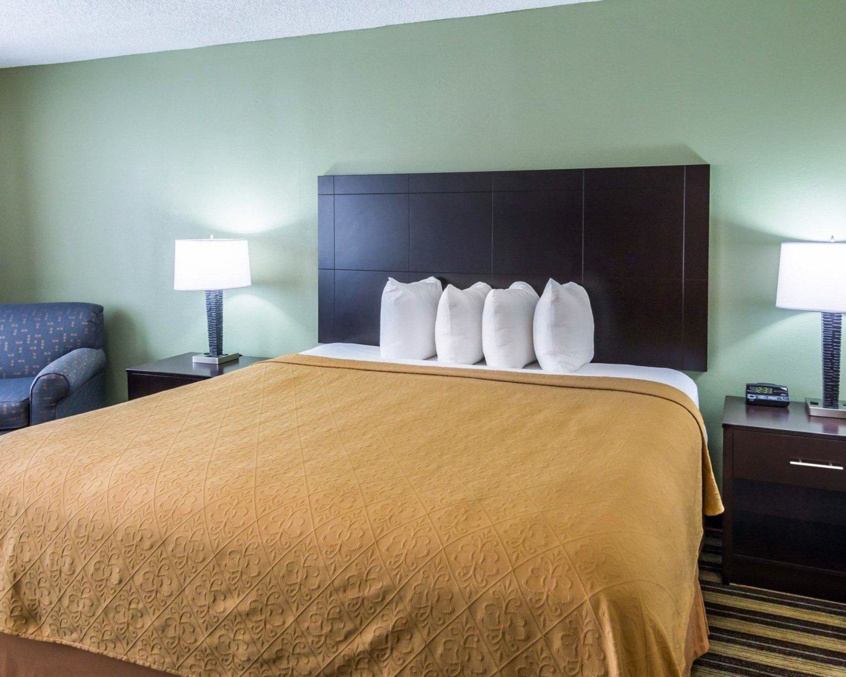 A hotel room with a large bed, a headboard, two bedside tables with lamps, a chair, and a carpeted floor.