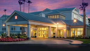 Un hotel de diseño moderno, con una entrada amplia con puertas de cristal, una zona cubierta con columnas y un jardín bien cuidado con plantas tropicales.