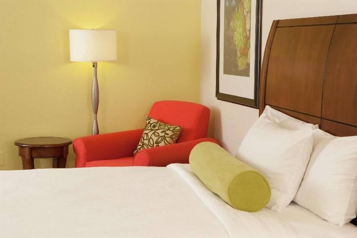 Une chambre d’hôtel avec un lit, un fauteuil rouge, une petite table, une lampe et un tableau encadré au mur.