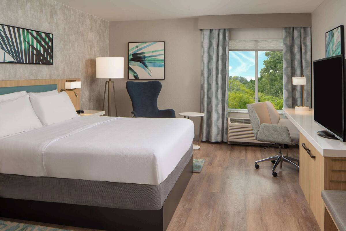 Une chambre d’hôtel avec un grand lit, un bureau avec un ordinateur, une chaise, une télévision et une fenêtre avec des rideaux.
