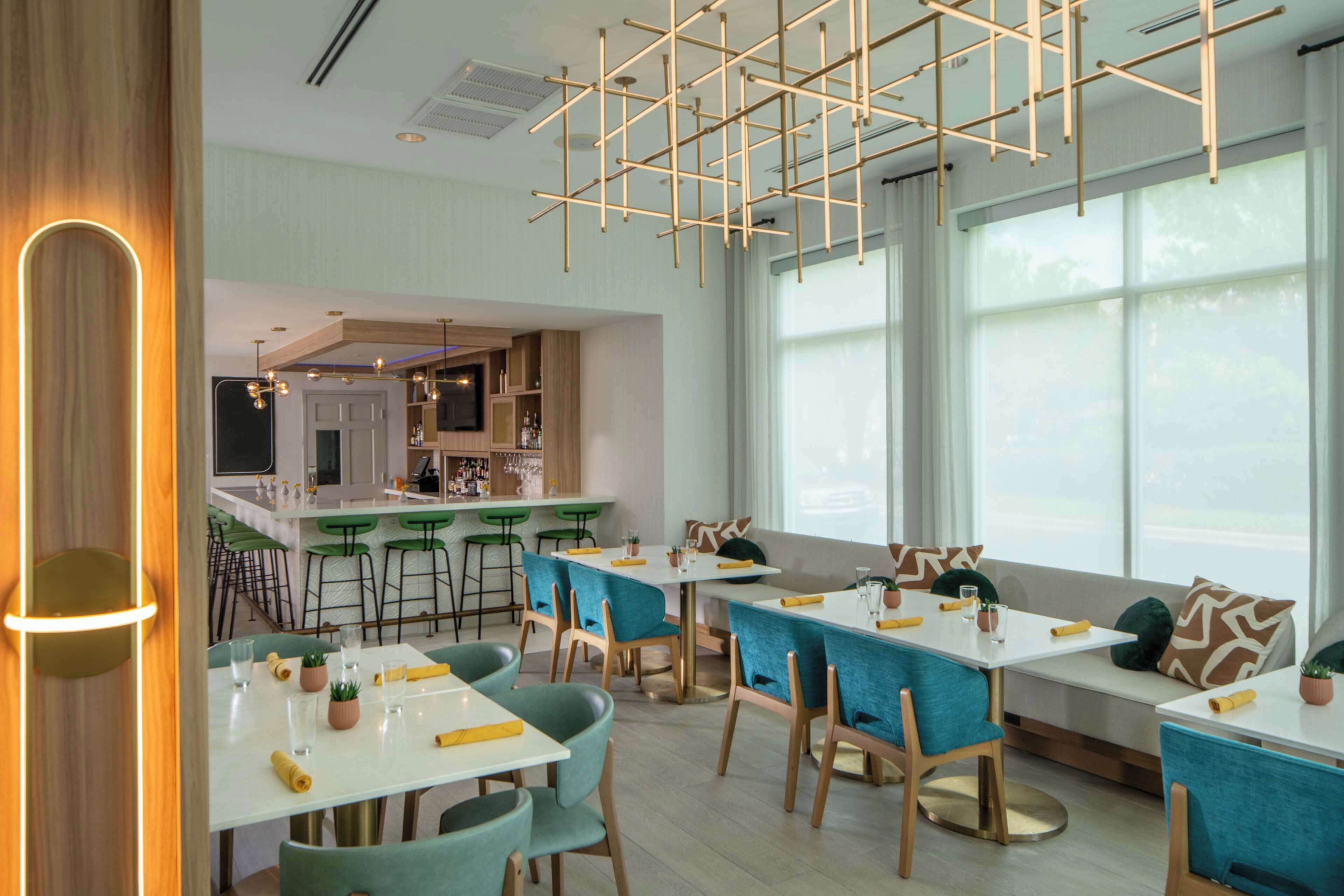 A modern dining area with tables and chairs, a kitchen in the background, and large windows.