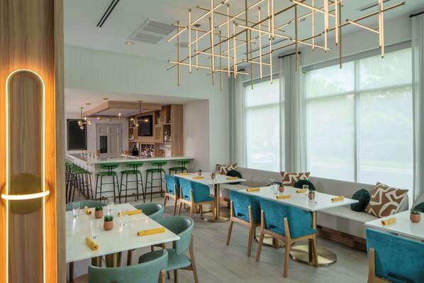 Un comedor moderno con mesas y sillas, una cocina al fondo y ventanas amplias.