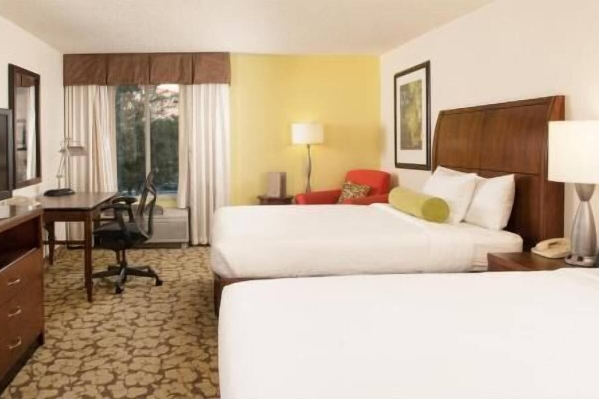 Une chambre d’hôtel avec deux lits, un bureau, une télévision, une chaise, une lampe et une fenêtre avec des rideaux.