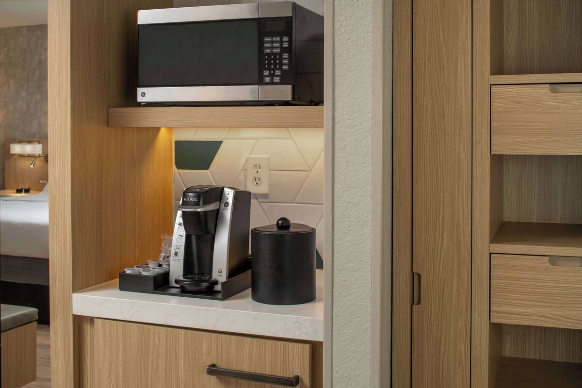 Une petite cuisine équipée d’un four à micro-ondes, d’une machine à café et d’un récipient noir posé sur un plan de travail.