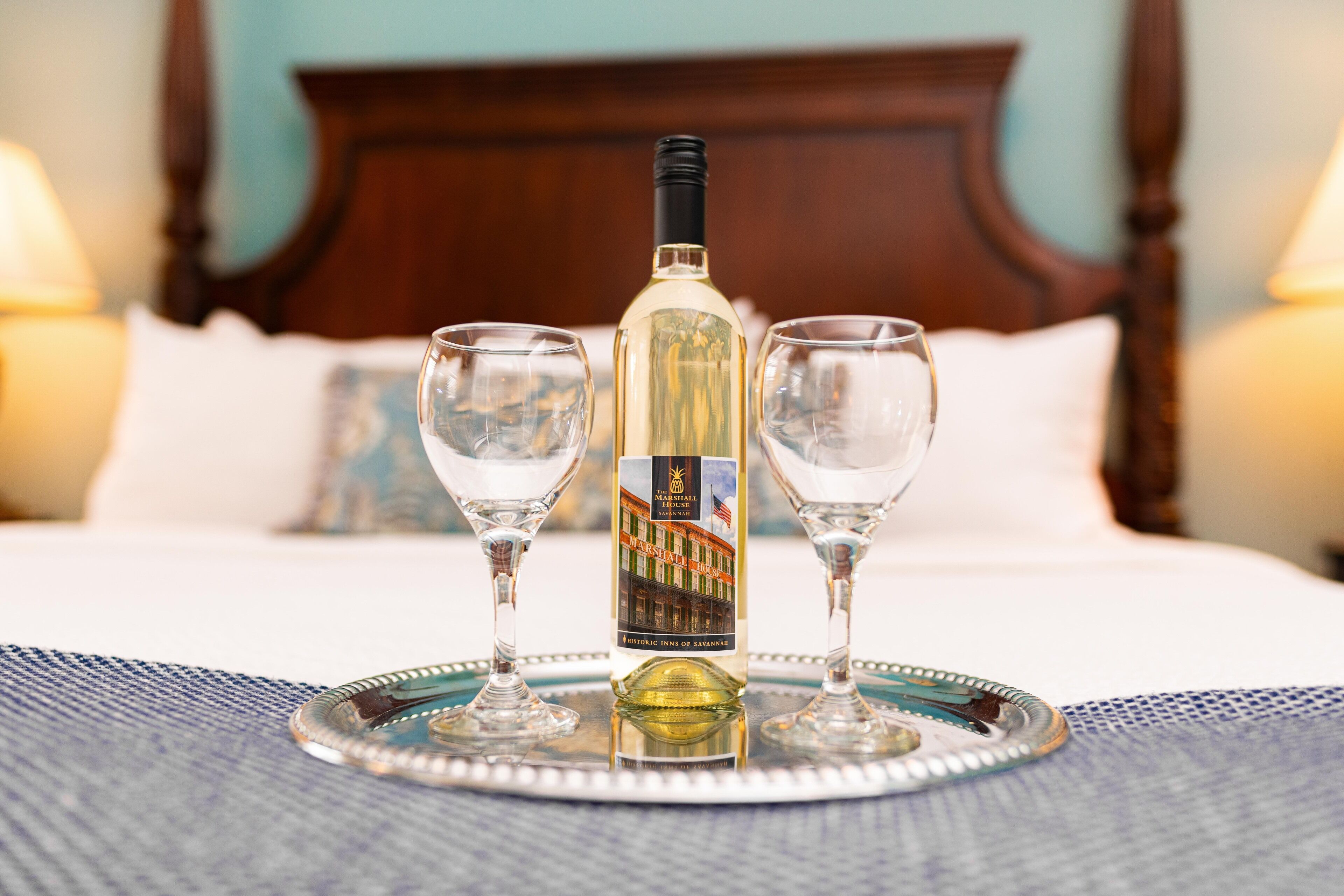 A bottle of wine on a tray with two glasses, set on a bed with a wooden headboard and pillows.
