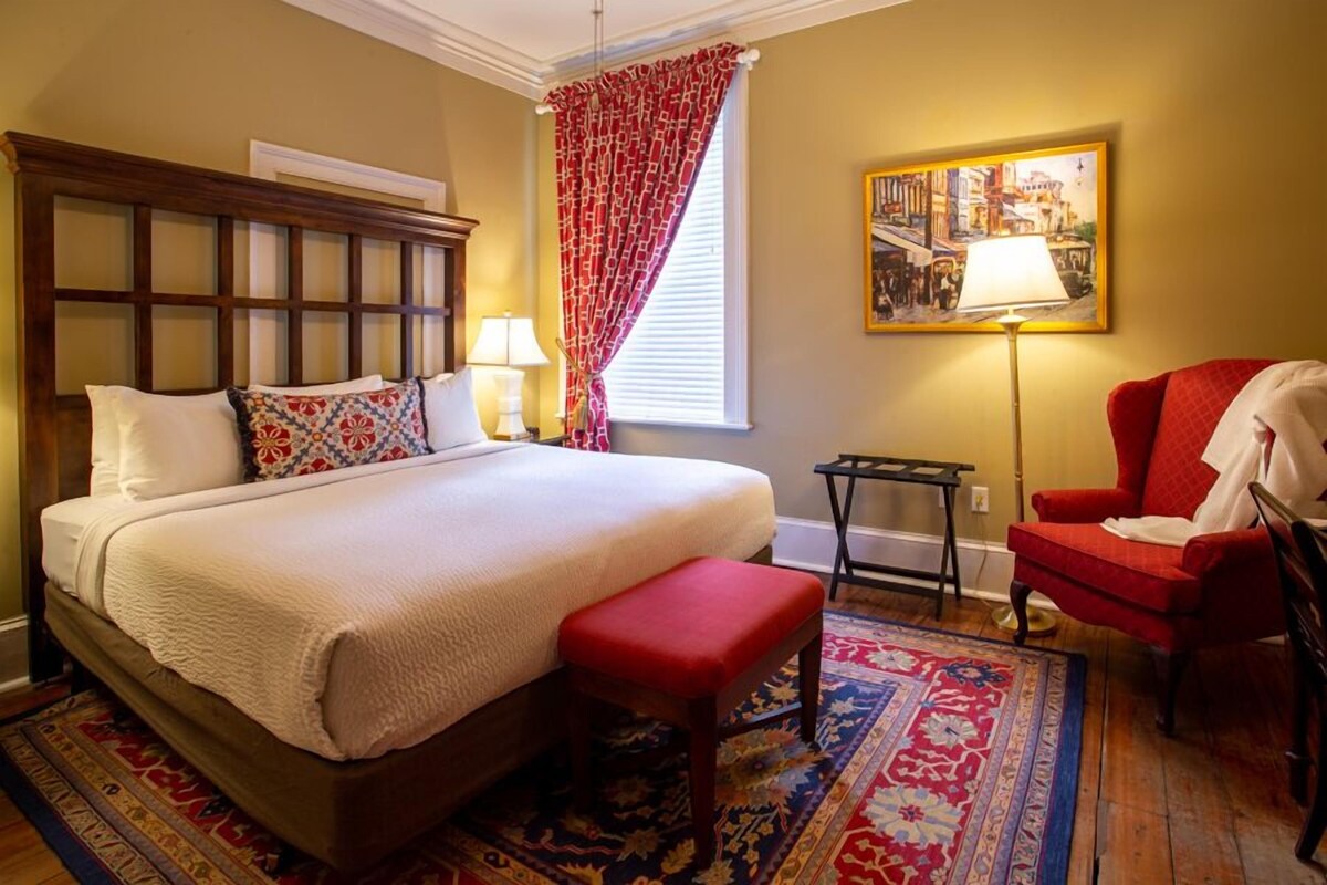 A bedroom with a large bed, a red chair, a small table, and a painting on the wall.