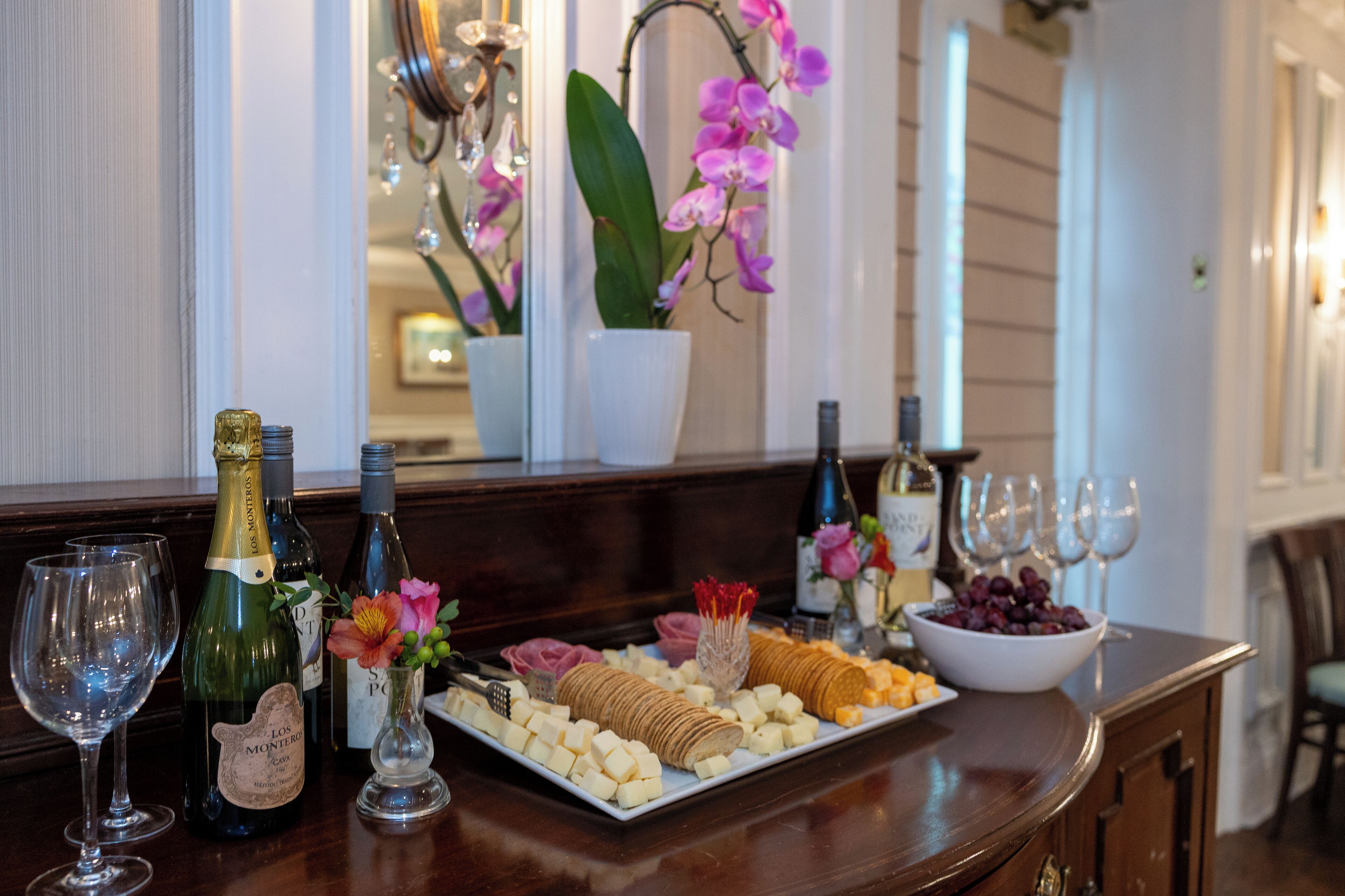 A bar area with a wine and cheese board, bottles, glasses, and orchids.