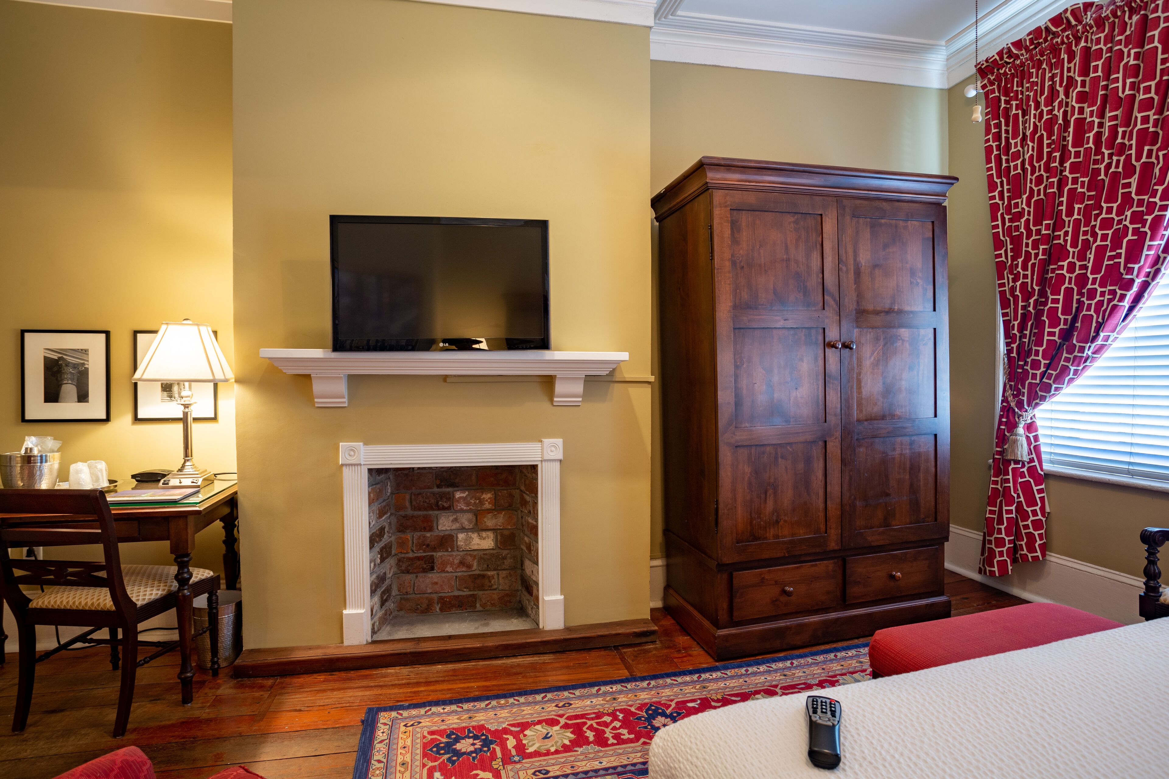 A hotel room with a fireplace, a large wooden cabinet, a television on a mantel, a desk with a lamp, and a bed with a red accent.