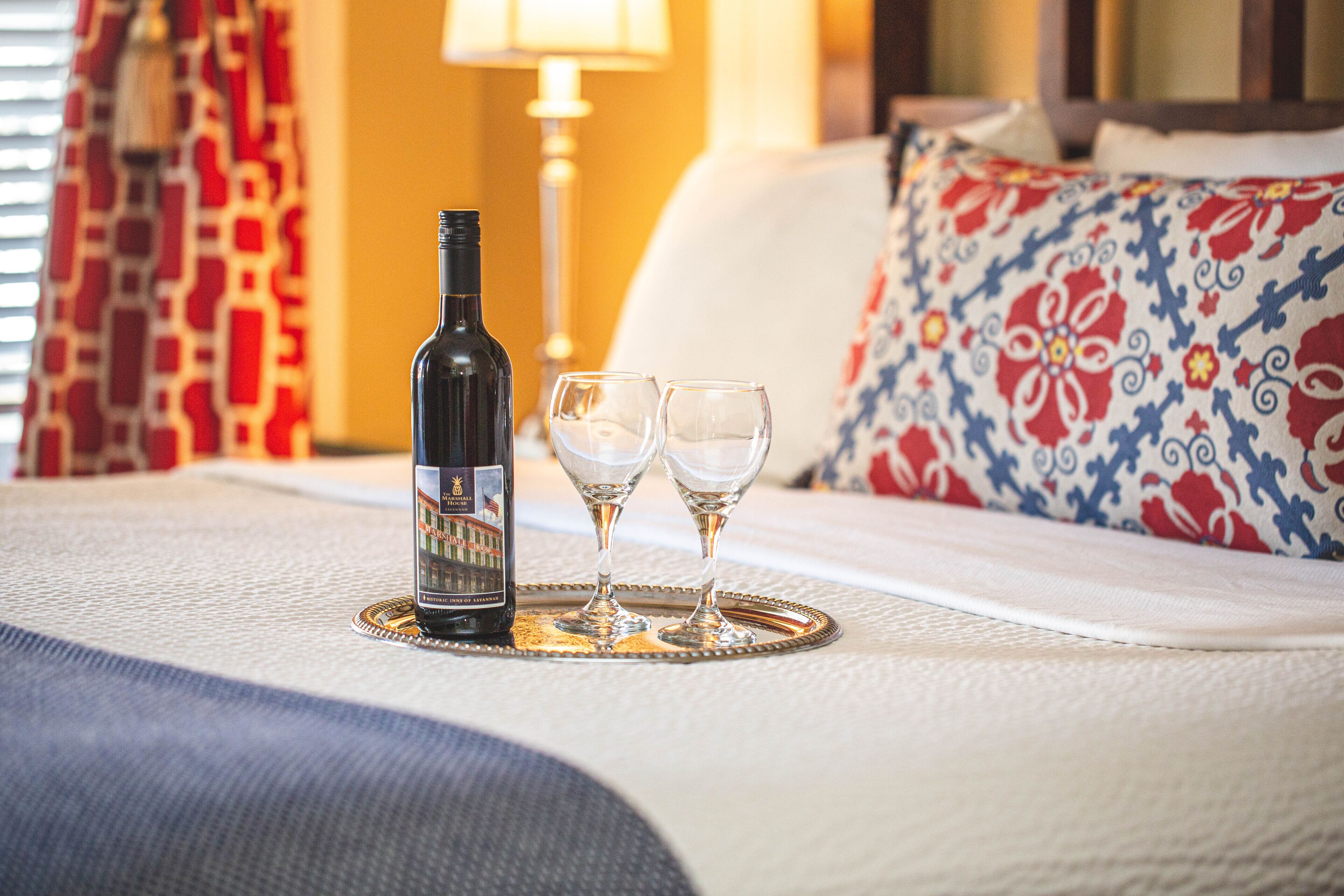 Una botella de vino y dos copas sobre una bandeja, sobre la cama, junto a una almohada estampada y una lámpara al fondo.