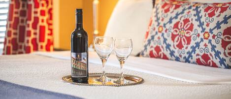 A bottle of wine and two glasses on a tray on a bed with a patterned pillow and a lamp in the background.