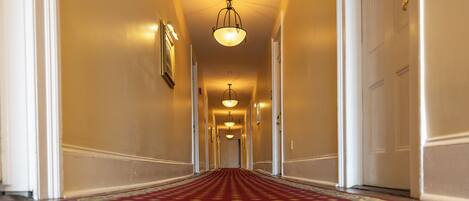 Un pasillo de hotel con alfombra roja, paredes blancas y varias puertas.