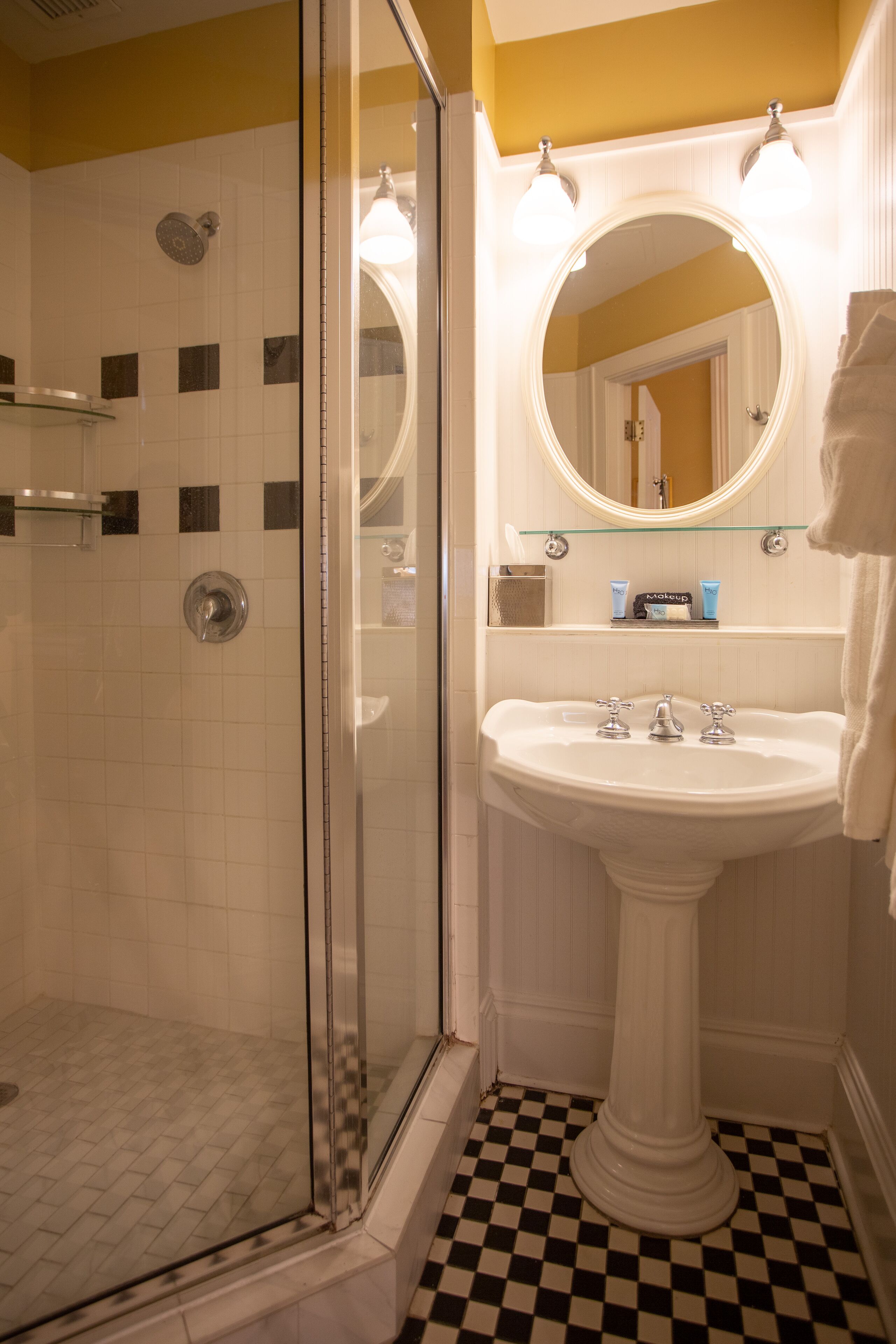 A bathroom with a glass-enclosed shower, a pedestal sink, and a mirror with lights.