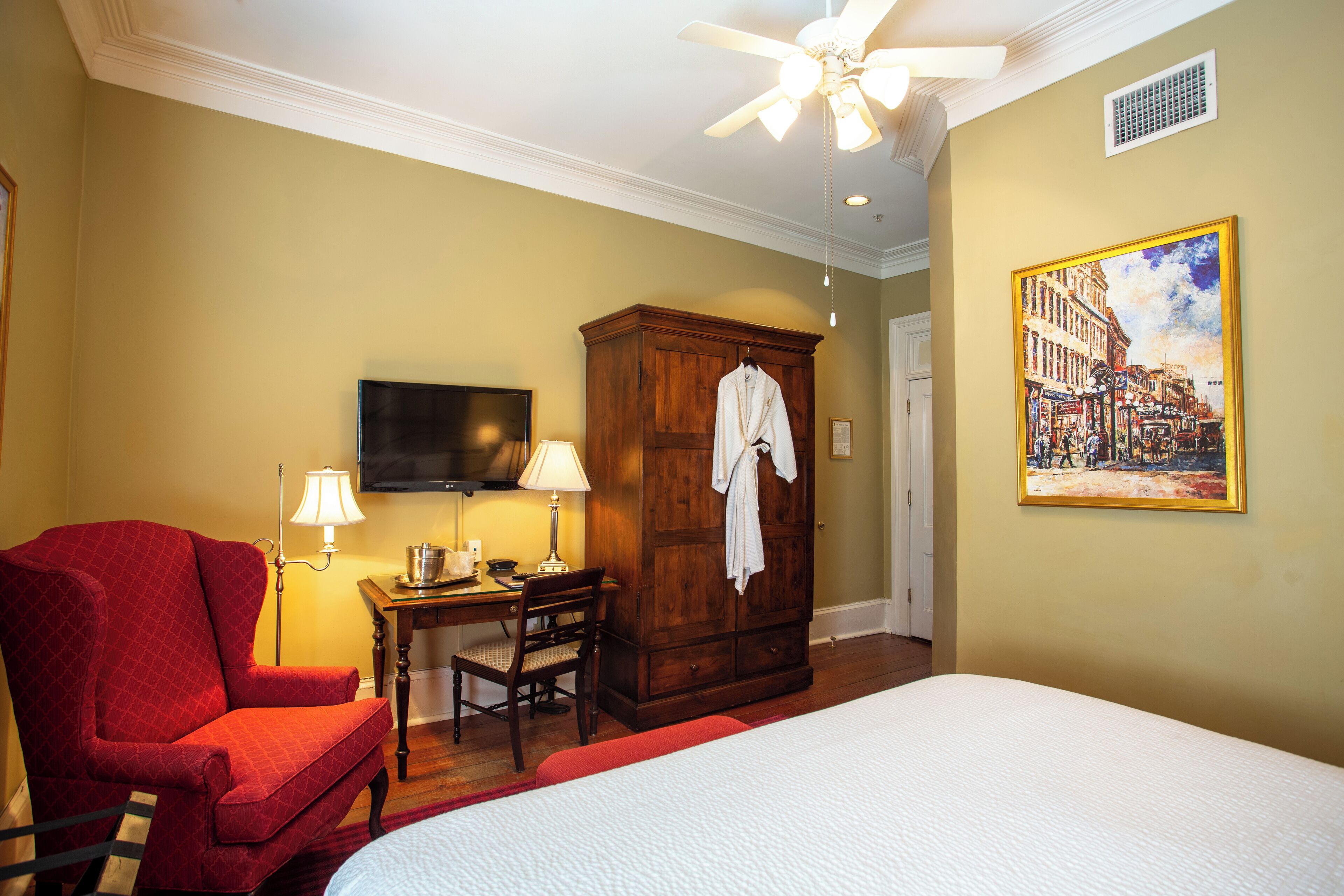 A hotel room with a bed, a red armchair, a wooden wardrobe, a desk with a lamp, and a painting on the wall.