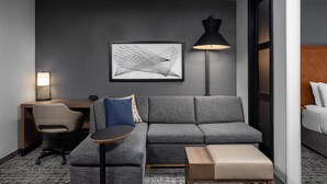 Una habitación de hotel moderna con un sofá gris, una mesita redonda pequeña, un escritorio con una silla, una lámpara y una obra de arte abstracta enmarcada en la pared.