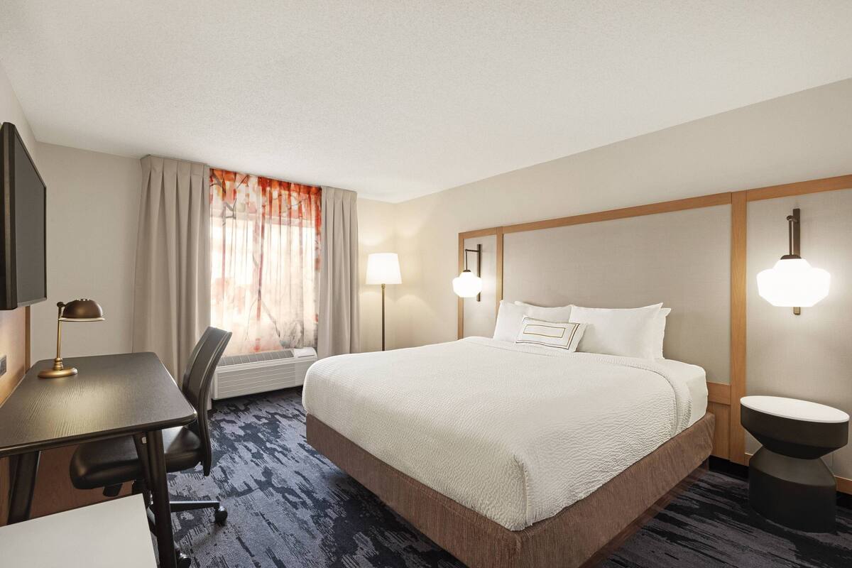 Habitación de hotel con una cama grande, un escritorio con lámpara, una silla, una mesita de noche y una ventana con cortinas.