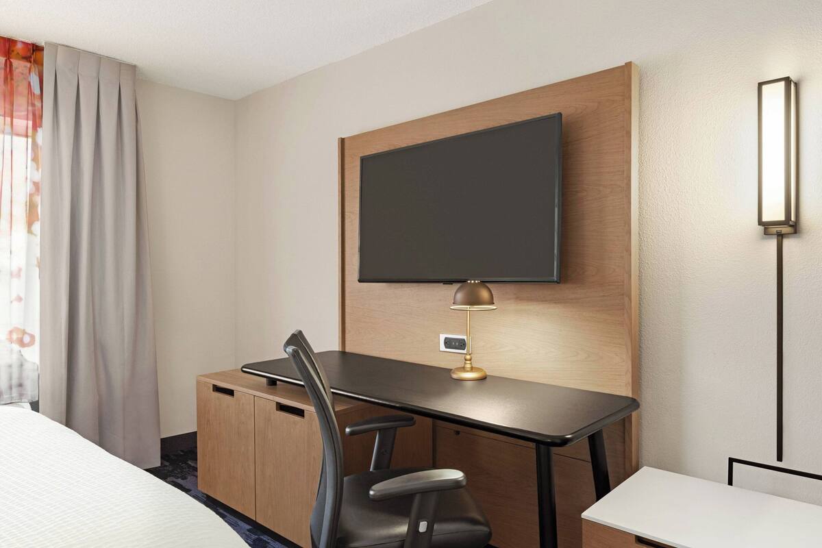 Una habitación de hotel con una cama, un escritorio con silla, un televisor montado en la pared y una mesita de noche.