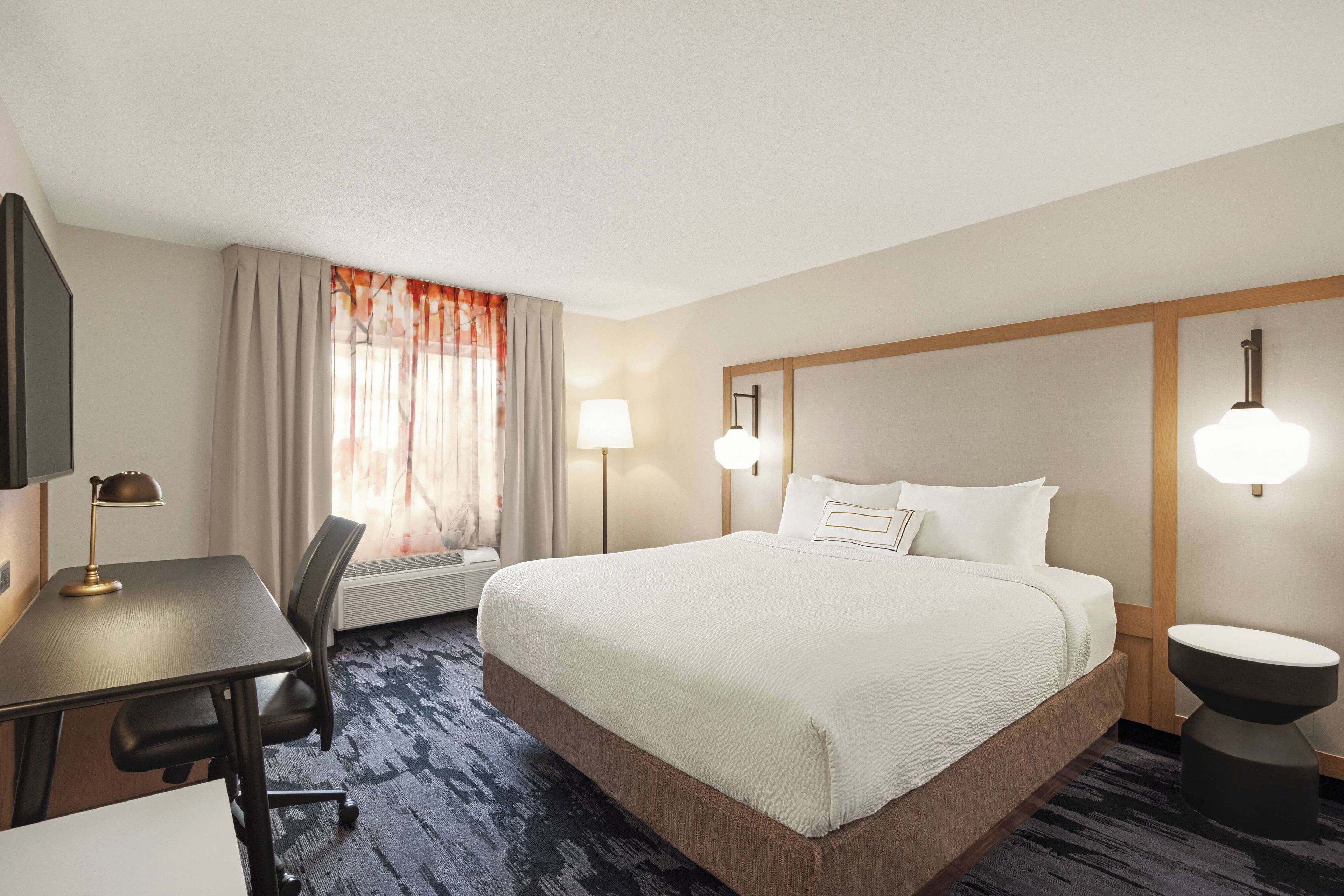 A hotel room with a large bed, a desk, a chair, a desk lamp, and a wall-mounted lamp.