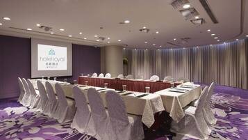 A conference room with a long table set for a meeting, equipped with chairs, water glasses, and a projector screen displaying a hotel logo.