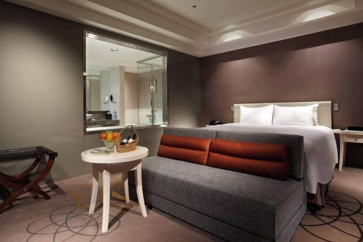 A hotel room with a bed, a small round table with a basket, a mirror, and a view of the bathroom.