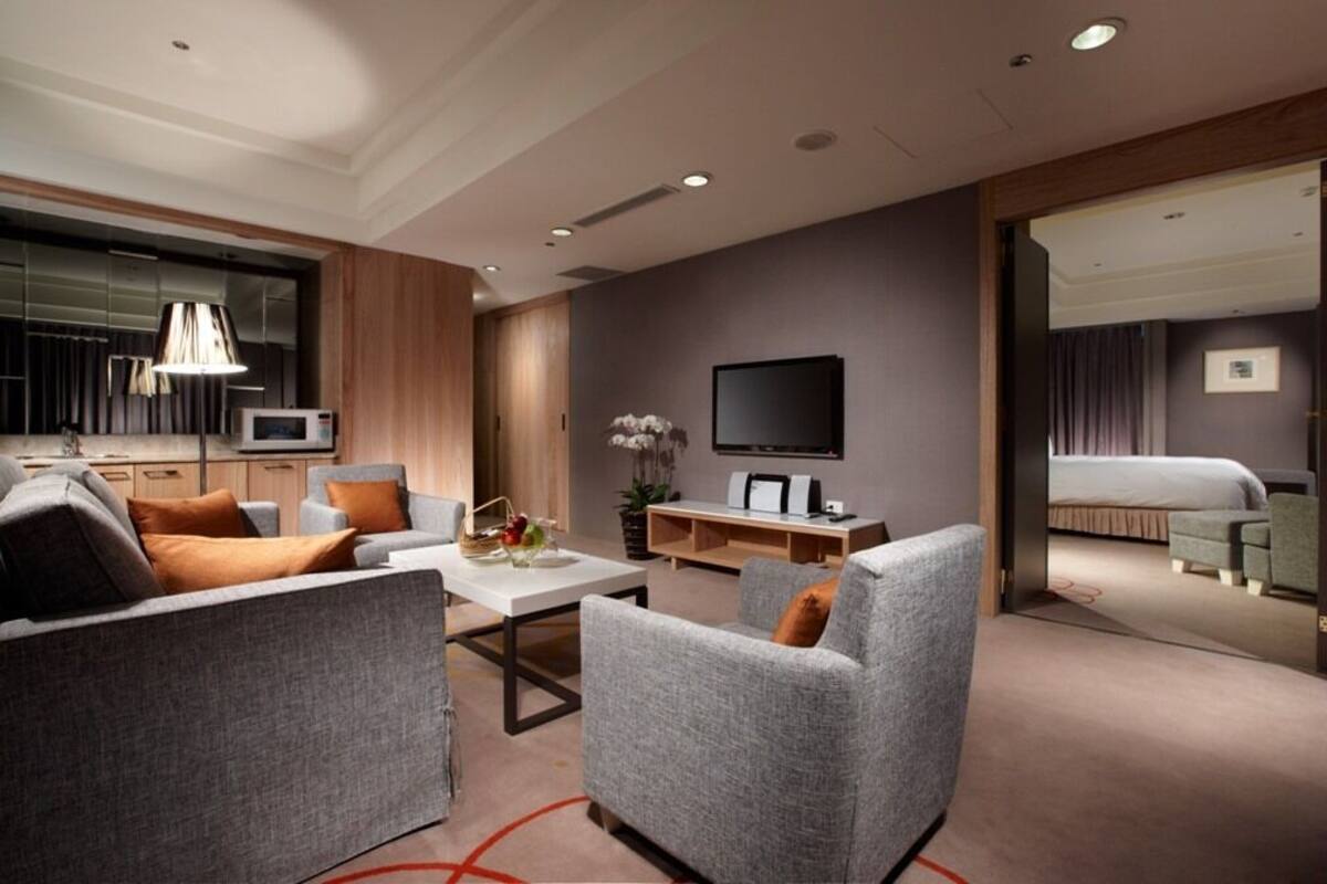 A modern hotel room with a living area, a flat-screen TV, a sofa, a coffee table, and a view into the bedroom.
