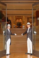 A person in a formal suit and white hat standing at a glass door, with a reflection visible in the glass.