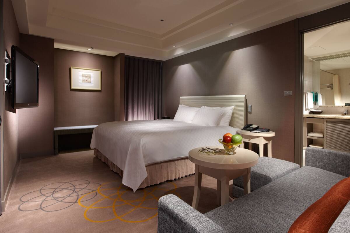A hotel room with a bed, a desk with a telephone, a chair, and a small table with fruit.
