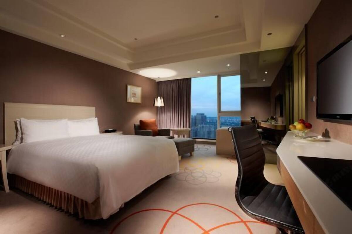 A hotel room with a large bed, a desk, a chair, a TV, and a city view.