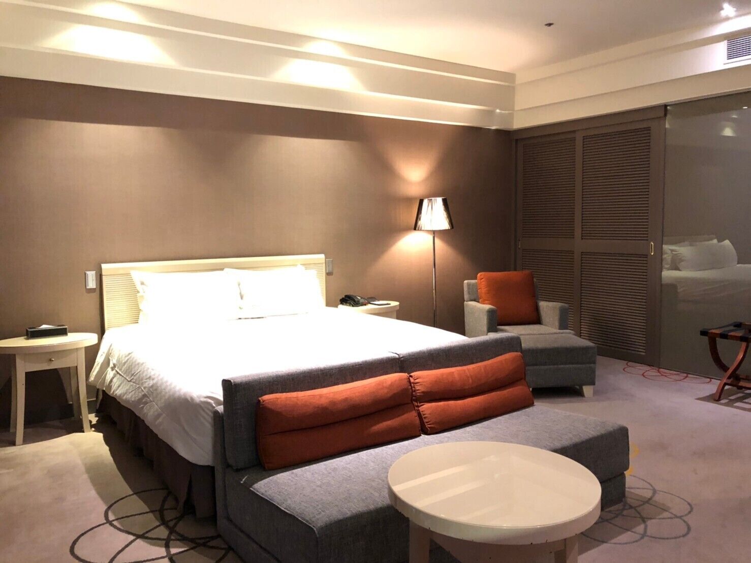 A hotel room with a large bed, a bedside table, a lamp, an armchair, a sofa, a small round table, and a sliding door closet.