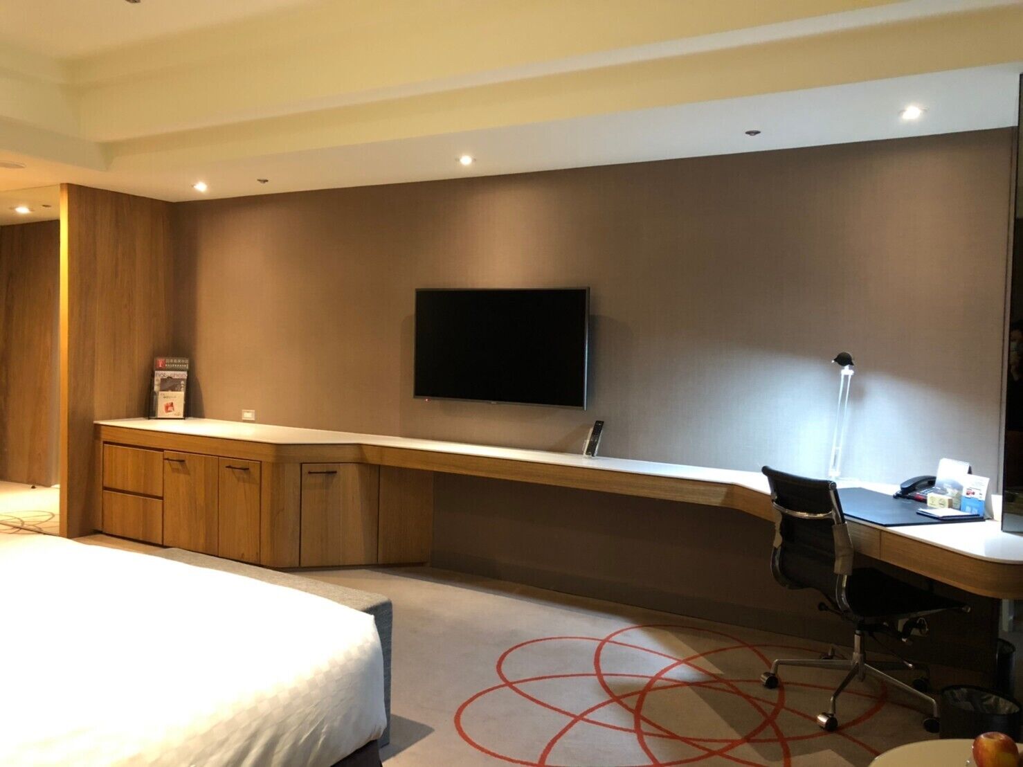 A hotel room with a bed, a desk with a chair, a flat-screen TV mounted on the wall, and a small table with a lamp.