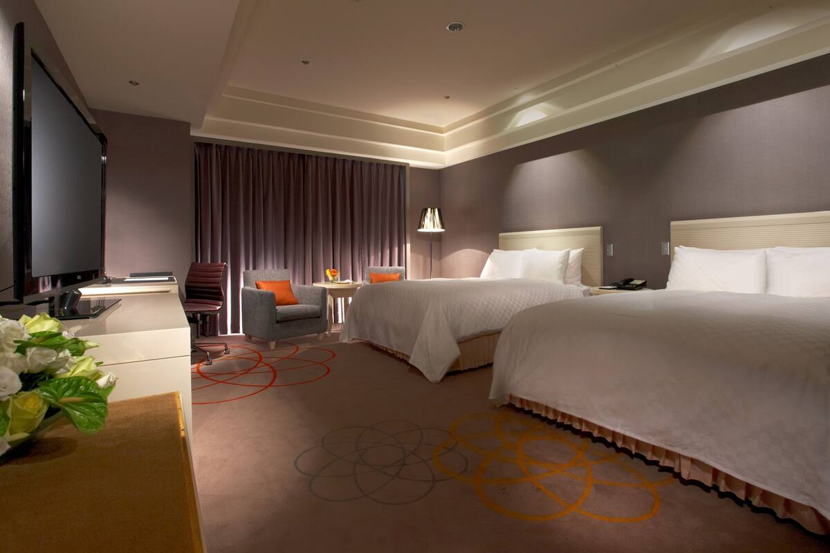 A hotel room with two beds, a desk, a chair, a TV, and a lamp.
