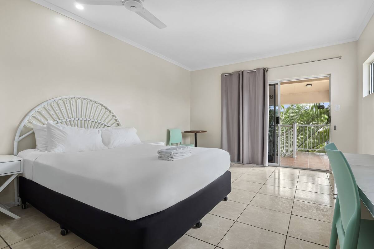 A hotel room with a bed, a desk, a chair, a ceiling fan, and a view of the outside.