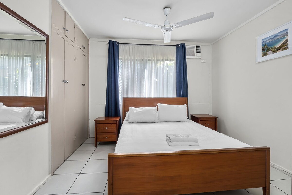 A bedroom with a wooden bed, a ceiling fan, a window with curtains, a bedside table, and a wardrobe.