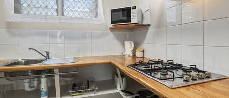 Una cocina con un lavamanos de acero inoxidable, un microondas, una estufa a gas y una ventana con cortina blanca.