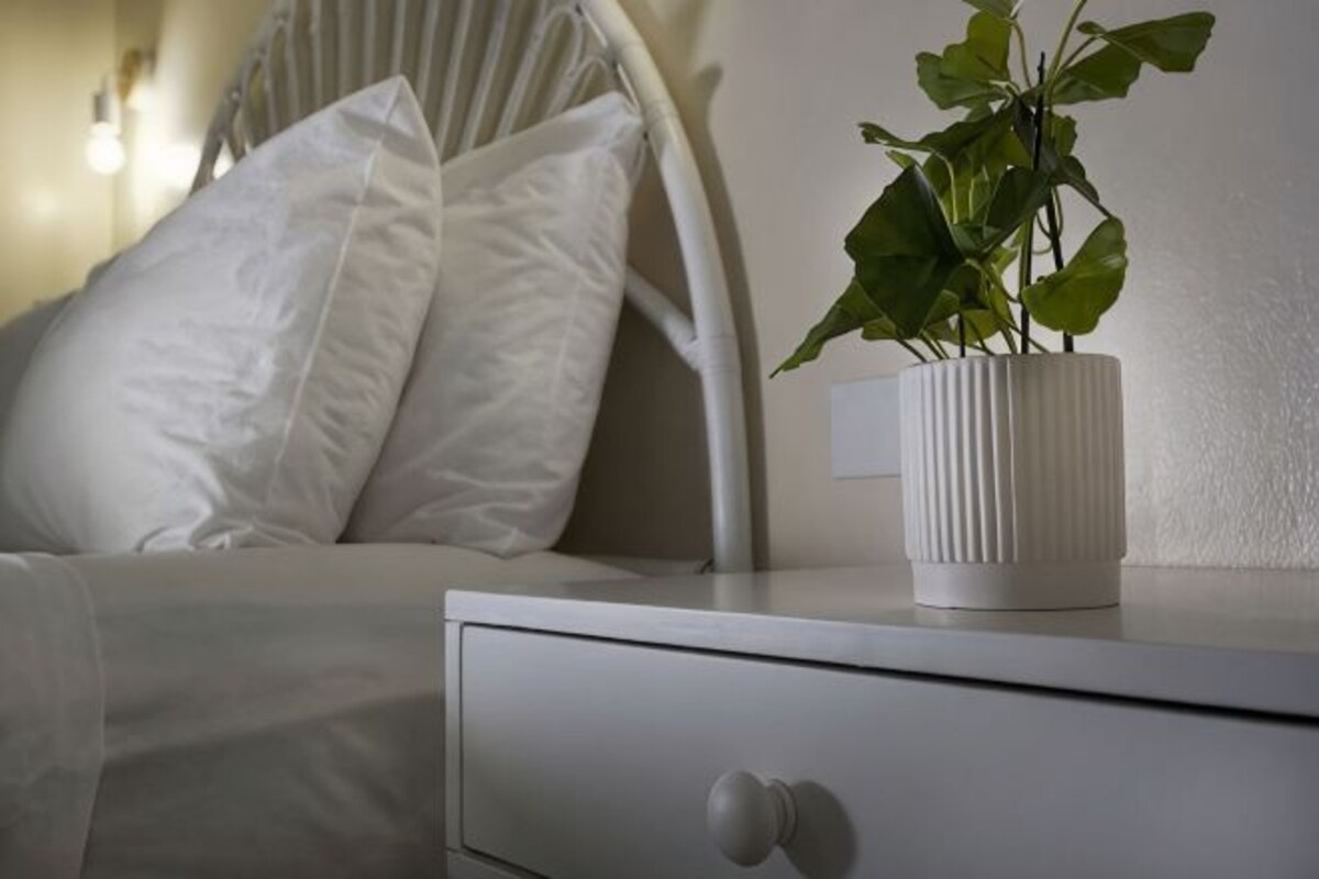 A white bedside table with a plant in a white pot, a white drawer, and a white bed with white pillows.
