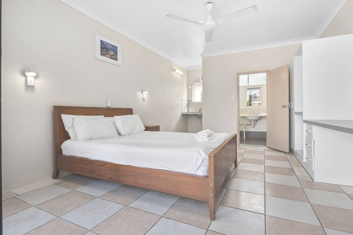 A bedroom with a wooden bed, white linens, a ceiling fan, and a bathroom visible through an open door.