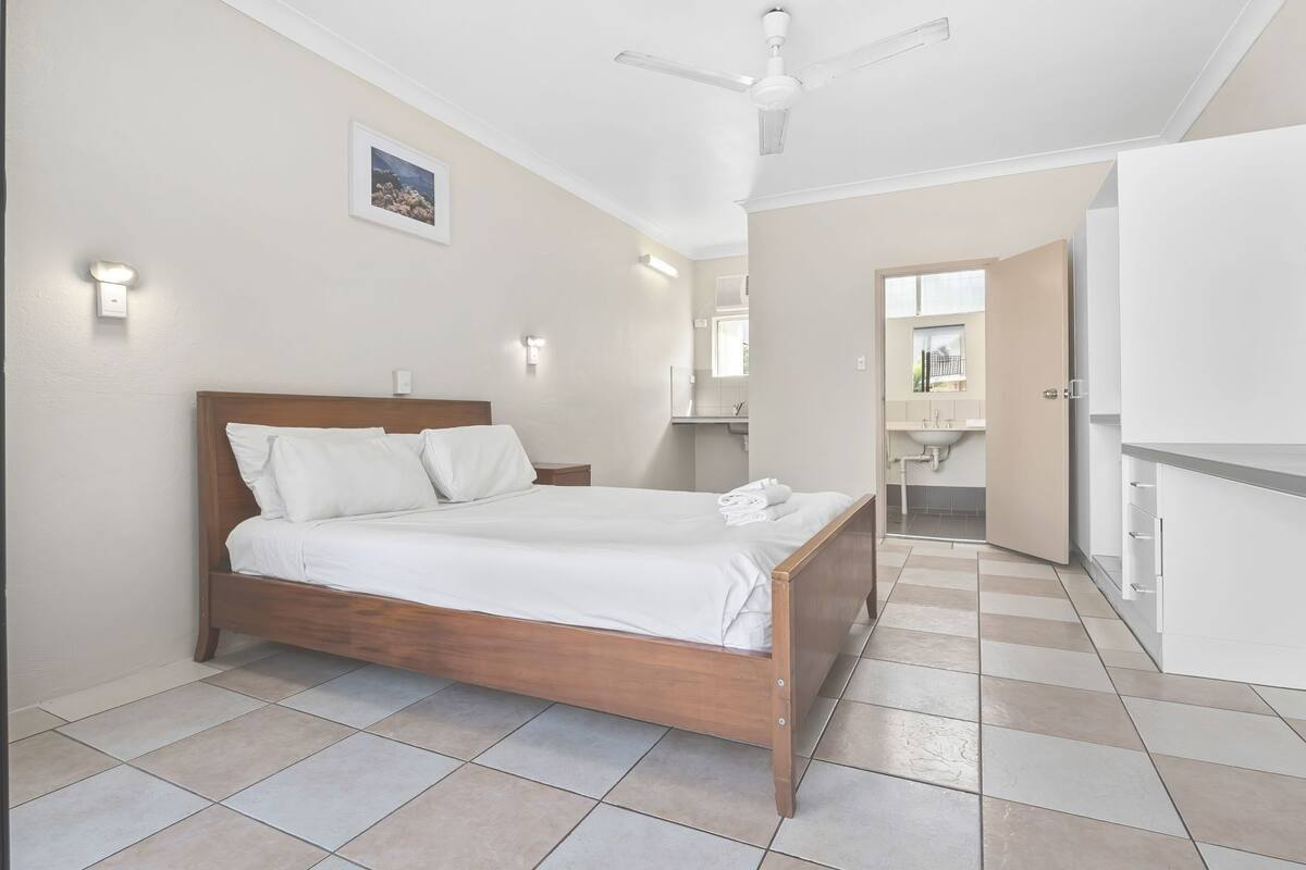 A bedroom with a wooden bed, a ceiling fan, a bathroom with a sink, and a hallway leading to a kitchen.