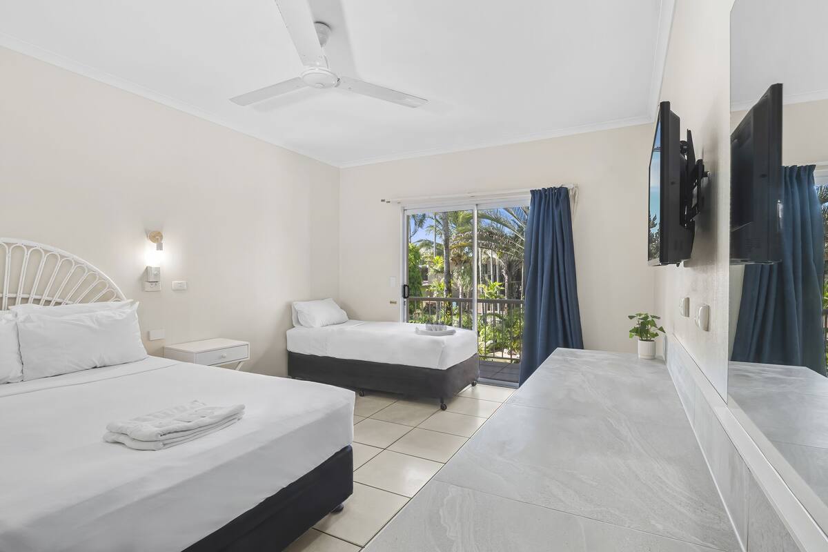 A hotel room with two beds, a ceiling fan, a TV, and a balcony with curtains.