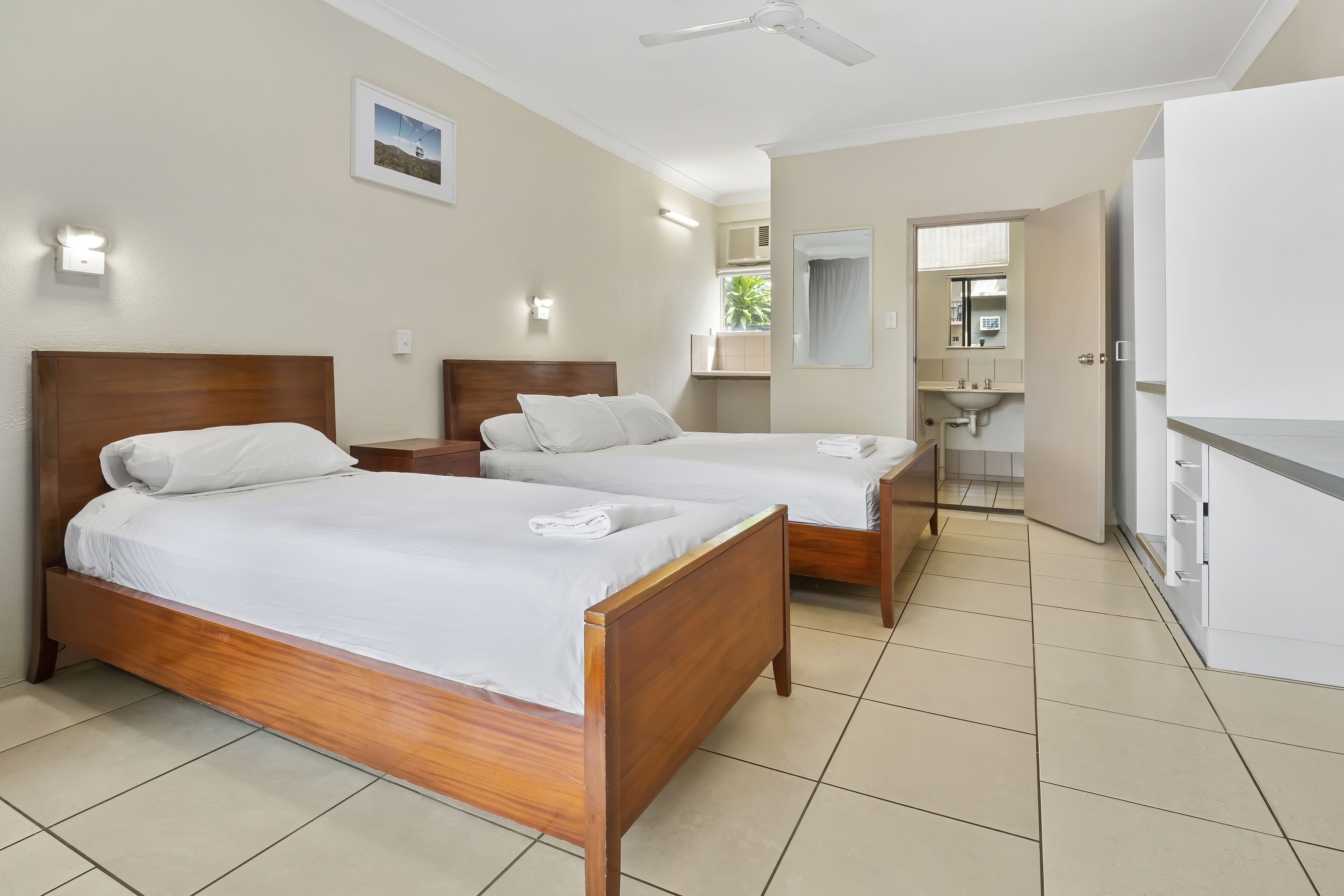 A hotel room with two wooden beds, a ceiling fan, a bathroom with a sink and mirror, and a small kitchenette.