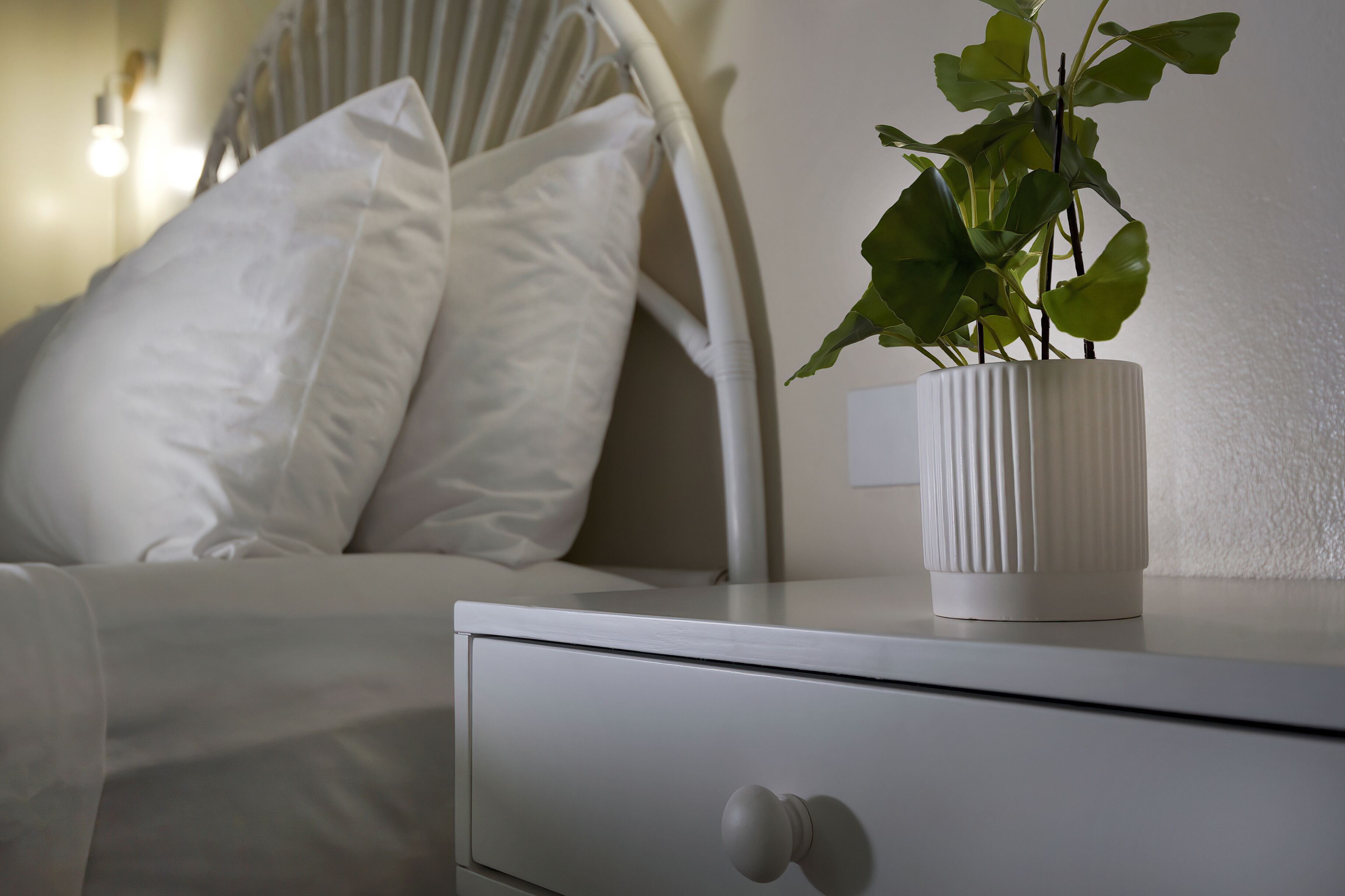 A bed with white bedding and pillows, a bedside table with a white vase containing a plant, and a white nightstand with a drawer.