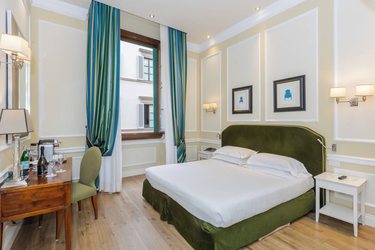 Ein Hotelzimmer mit einem großen Bett, einem Schreibtisch mit Stuhl, einem kleinen Tisch und einem Fenster mit Vorhängen.