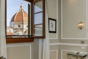 A room with a window view of a historic building, a framed picture on the wall, and a wall-mounted light fixture.